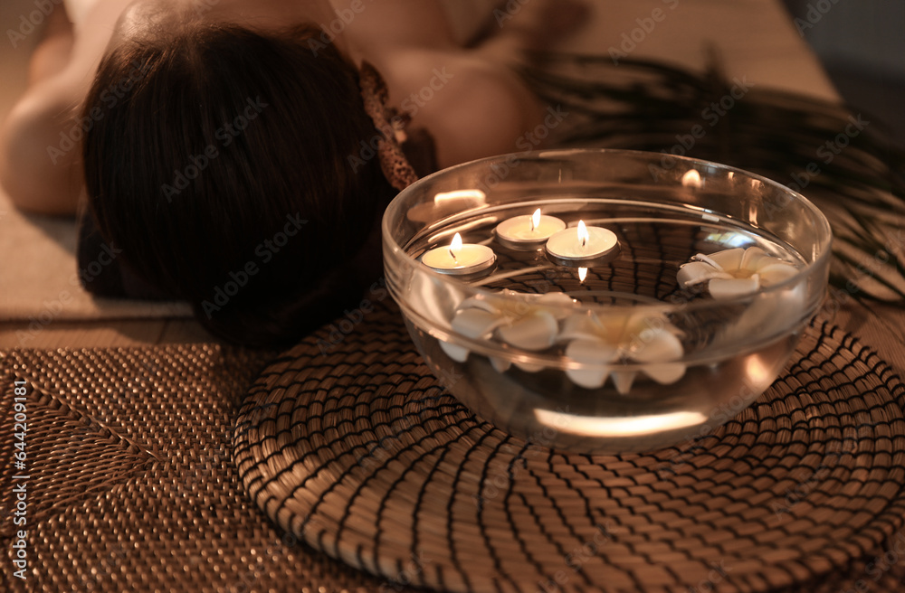 Bowl of water with burning candles and plumeria flowers on table in dark spa salon, closeup