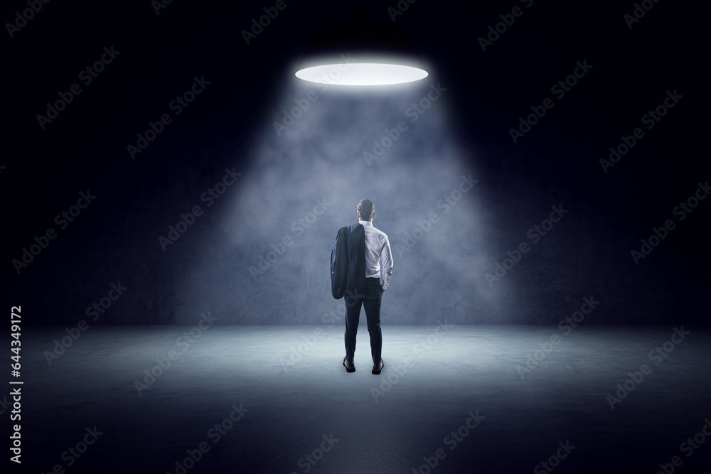 Back view of young man standing in spotlight of lamp in concrete interior.