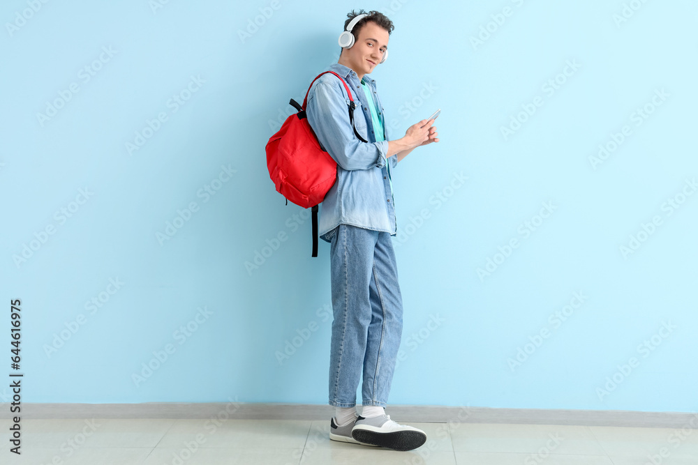 Male student with backpack and mobile phone listening music in headphones near blue wall