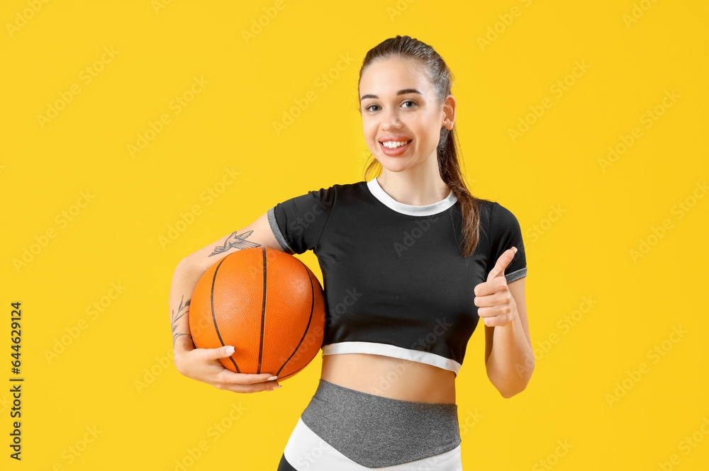 Sporty young woman with ball showing thumb-up on yellow background