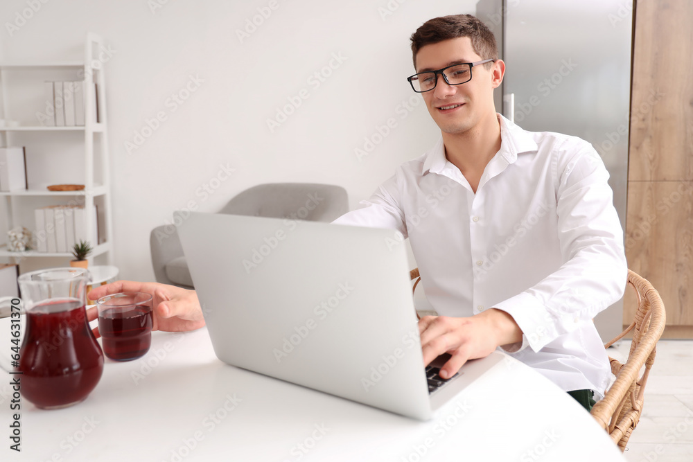 Young man with laptop taking glass of juice in kitchen