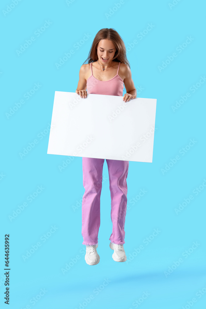 Young woman with blank poster jumping on blue background