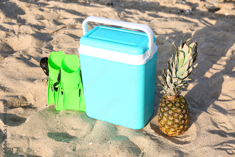 Beach cooler, flippers and ripe pineapple on sand