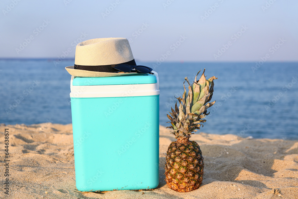 Modern beach cooler, accessories and ripe pineapple on sand