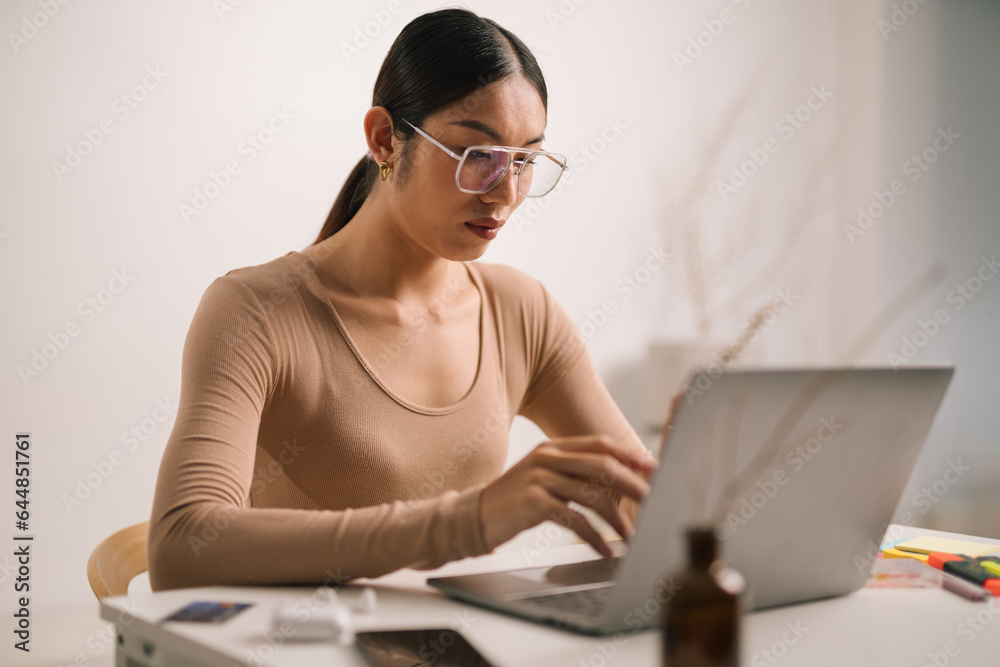 Asian male in eyeglasses and earphones using laptop computer while sitting at home office desk. LGBT