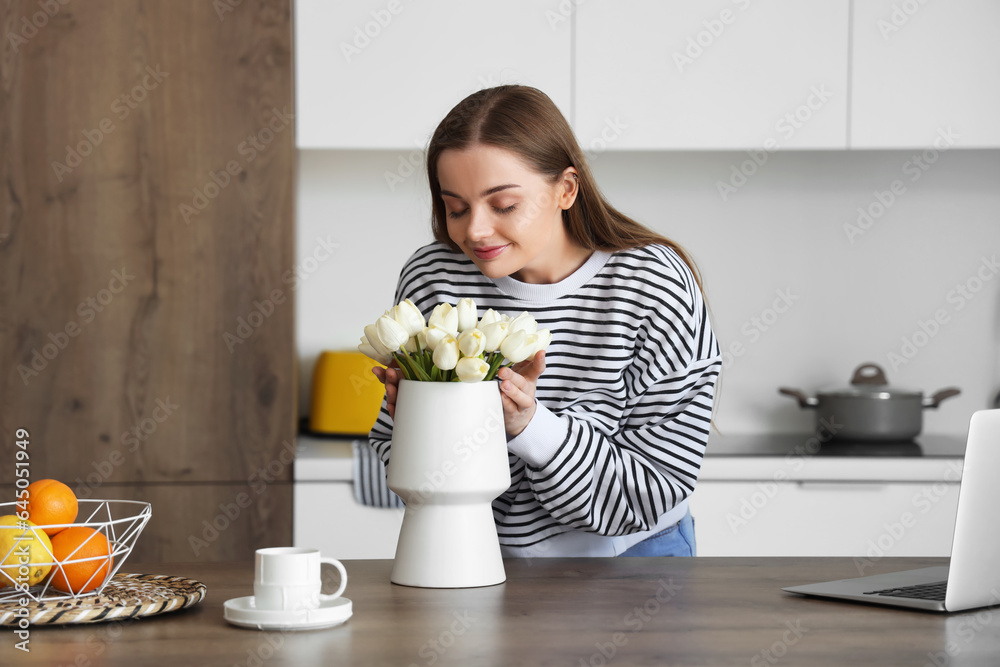 Beautiful young woman holding vase with white tulip flowers at table in kitchen