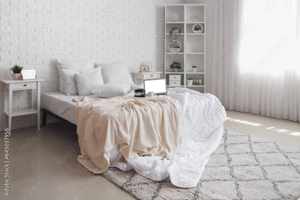 Messy bed with white blanket, coffee on tray and modern laptop in light bedroom