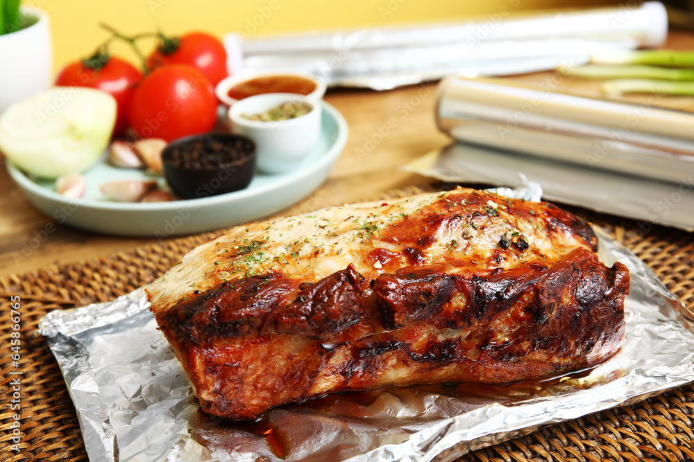 Aluminium foil with tasty baked meat, vegetables and spices on wooden table in kitchen, closeup