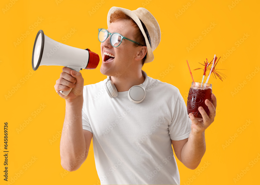 Young man with glass of juice shouting into megaphone on yellow background