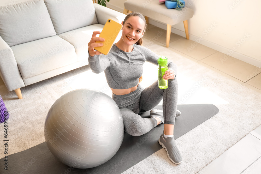 Sporty young woman with water bottle taking selfie at home