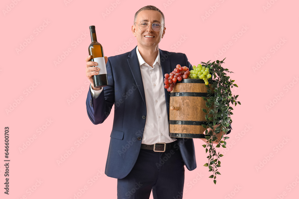 Mature man with bottle of wine, barrel and grapes on pink background