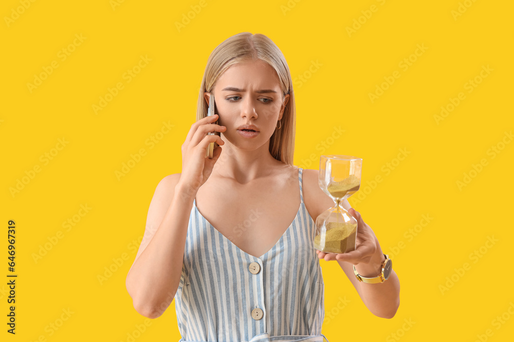 Stressed young woman with hourglass talking by mobile phone on yellow background. Deadline concept