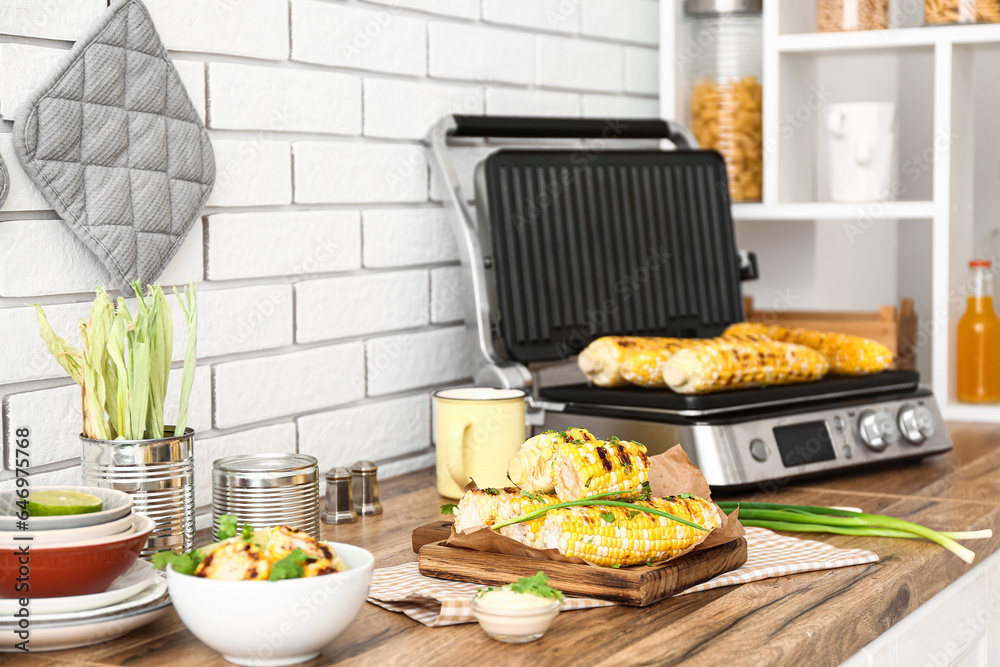 Board and electric grill with tasty corn on wooden kitchen counter