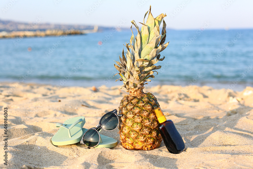 Stylish black sunglasses with pineapple, sunscreen and flip flops on seashore