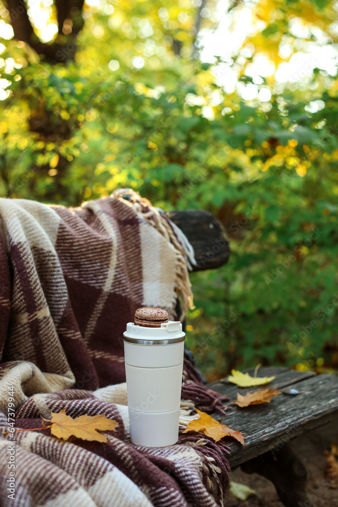 Cup of coffee with macaroon, fallen leaves and plaid on bench in autumn park