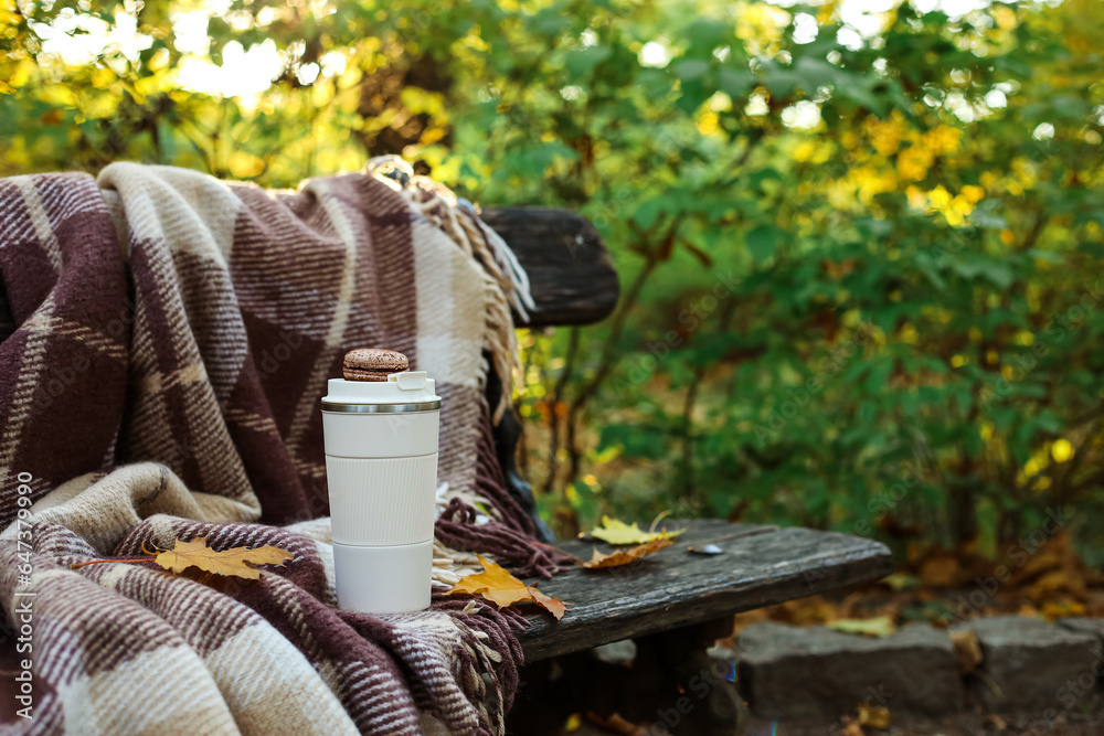 Cup of coffee with macaroon, fallen leaves and plaid on bench in autumn park