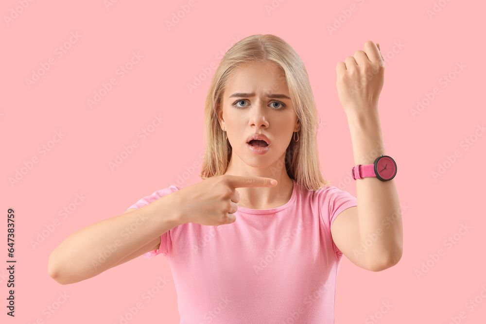 Stressed young woman pointing at wristwatch on pink background. Deadline concept