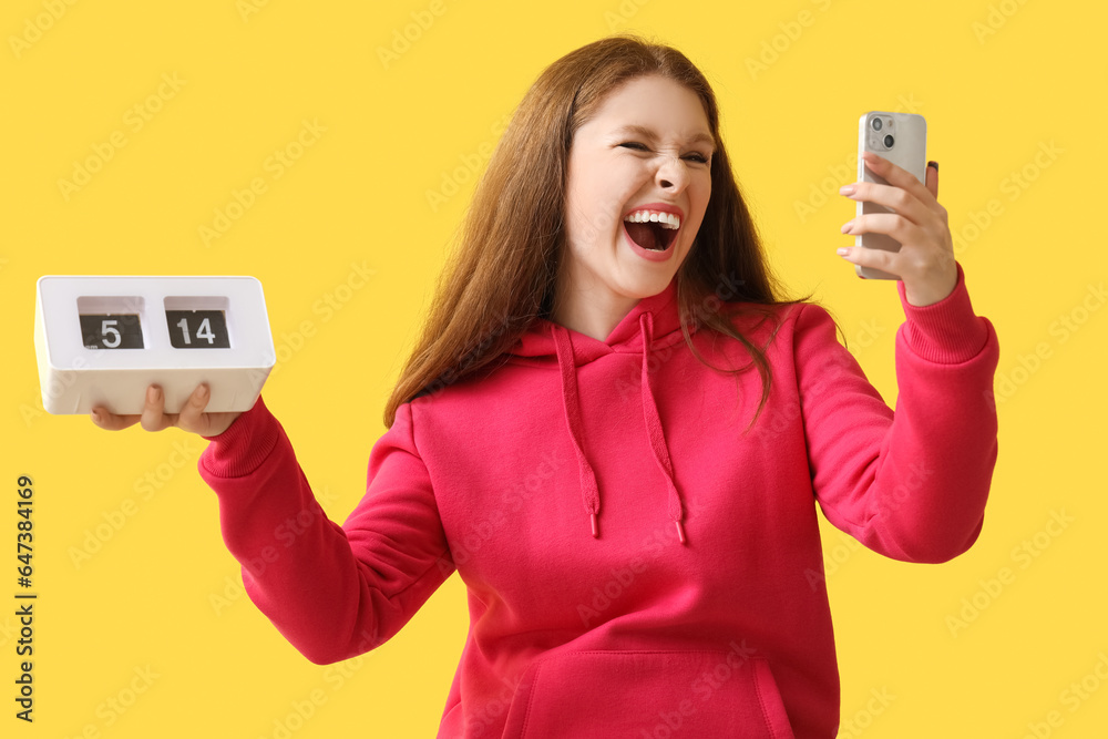 Stressed young woman with clock and mobile phone on yellow background. Deadline concept