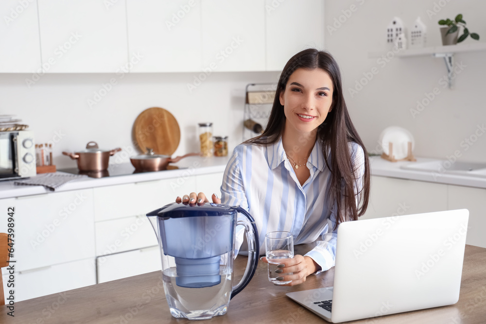 Young woman with water filter jug and laptop in kitchen