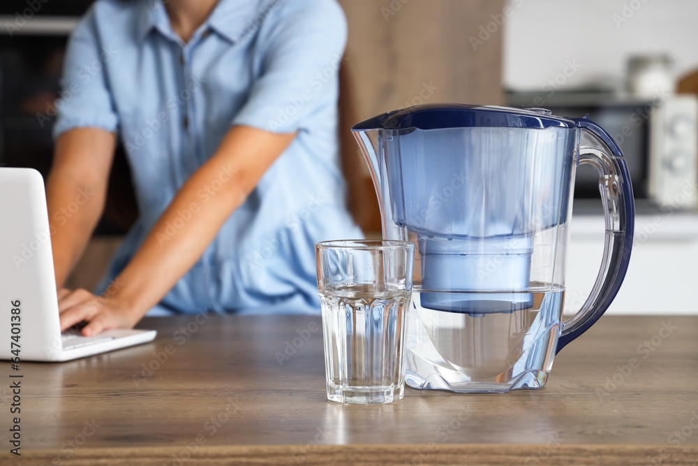 Beautiful young woman with water filter jug and glass using laptop in kitchen