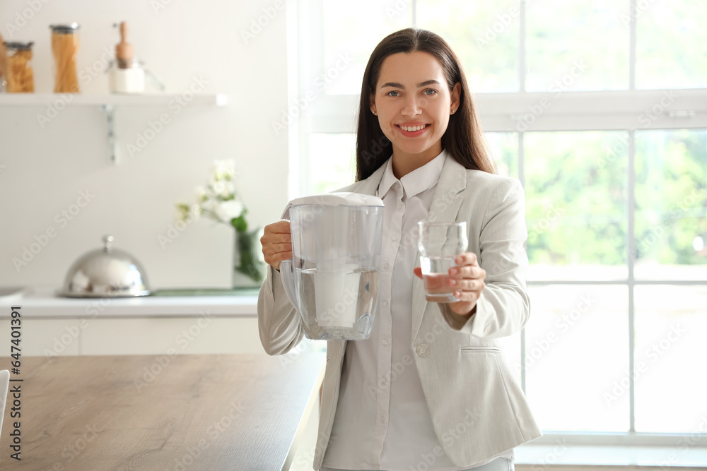 Beautiful young woman with water filter jug and glass in kitchen