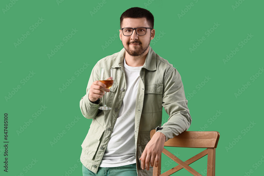 Young sommelier with glass of wine leaning on wooden chair against green background