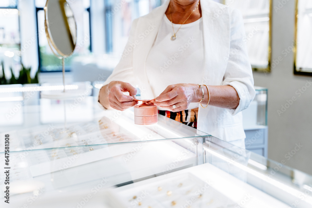 Close up of senior woman hands holding small gift with ribbon at couner in jewellery shop. Holidays 