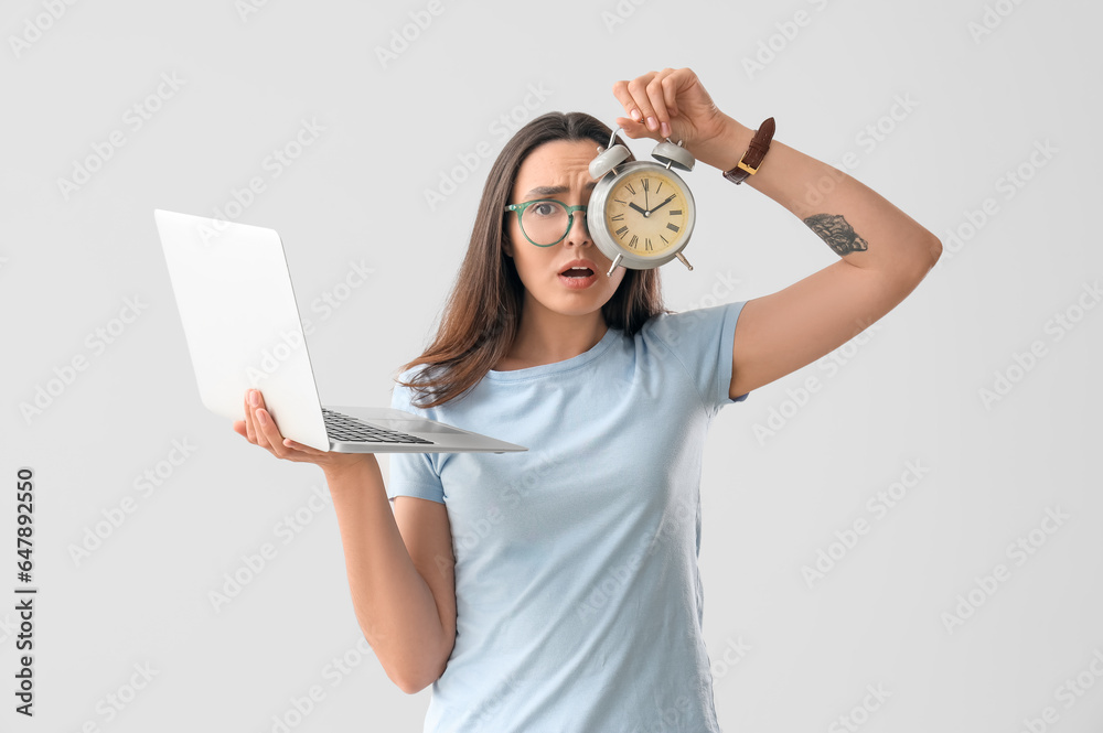 Stressed young woman with alarm clock and laptop on light background. Deadline concept