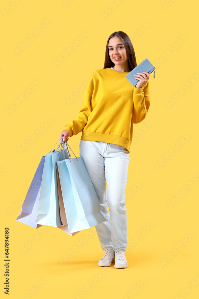 Young woman with wallet and shopping bags on yellow background