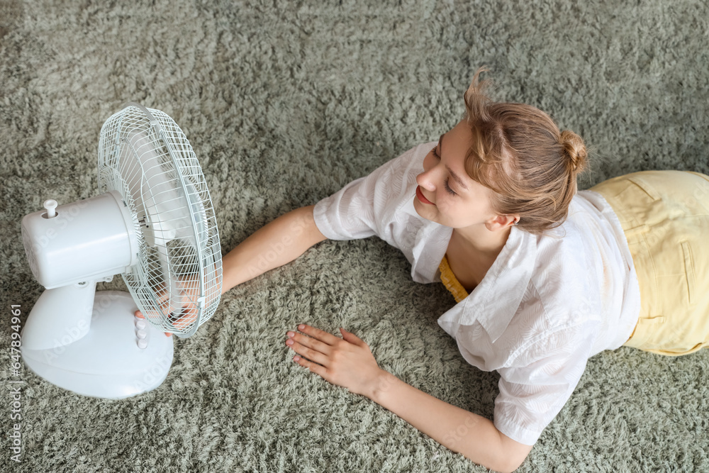 Young woman with blowing electric fan lying on floor at home