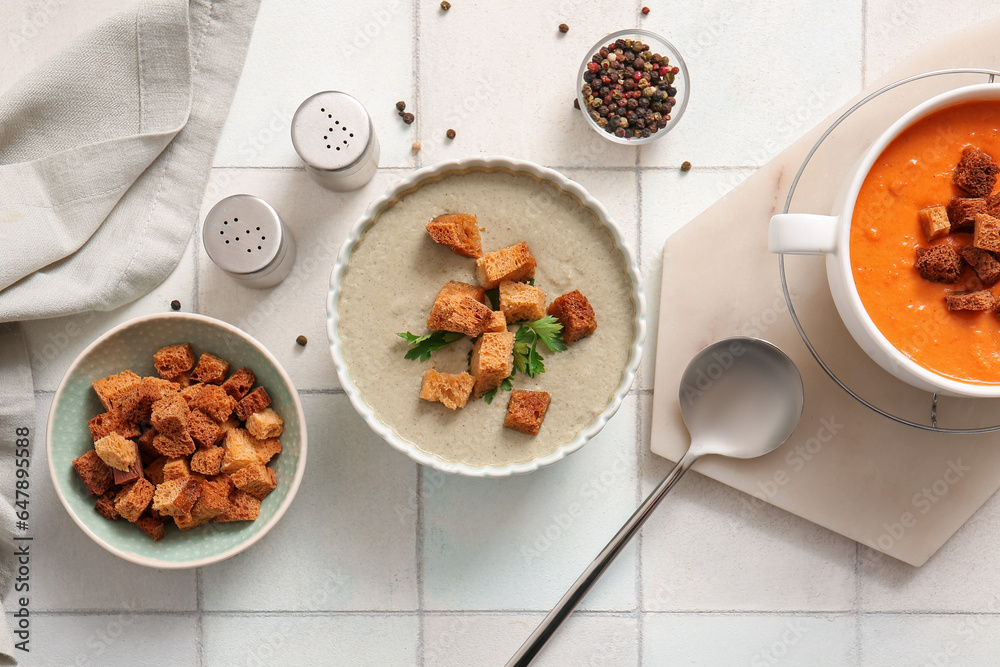 Pot and bowl of tasty cream soup with croutons on white tile background