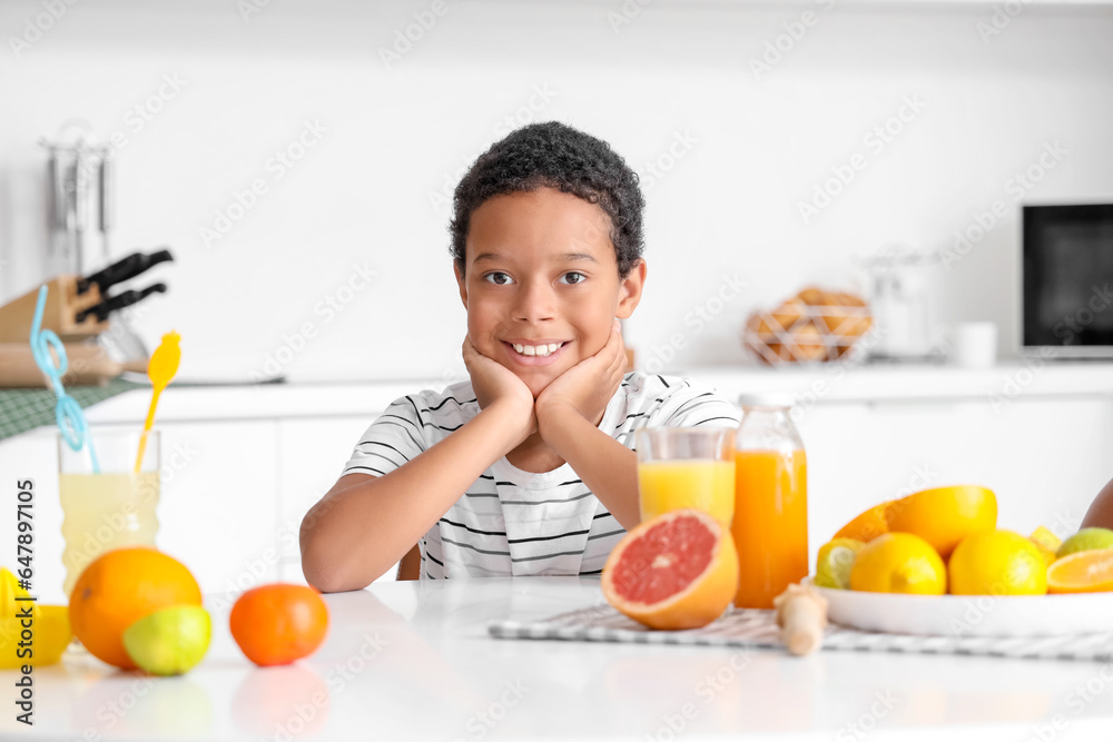 Little African-American boy with fresh juice and plate of different citruses sitting at table in kit