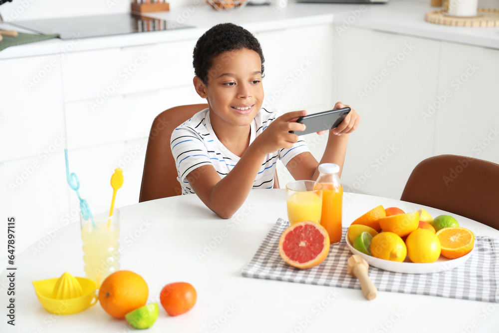 Little African-American boy with mobile phone, fresh juice and plate of different citruses in kitche