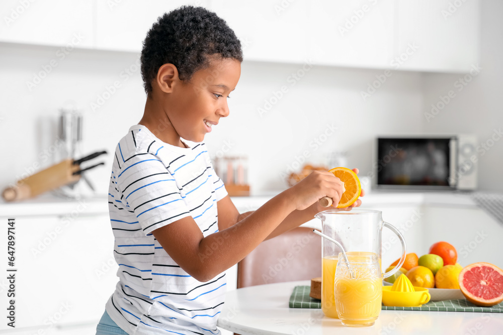 Little African-American boy making fresh citrus juice at table in kitchen