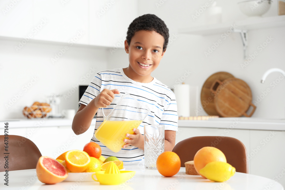 Little African-American boy pouring fresh citrus juice into glass from jug at table in kitchen