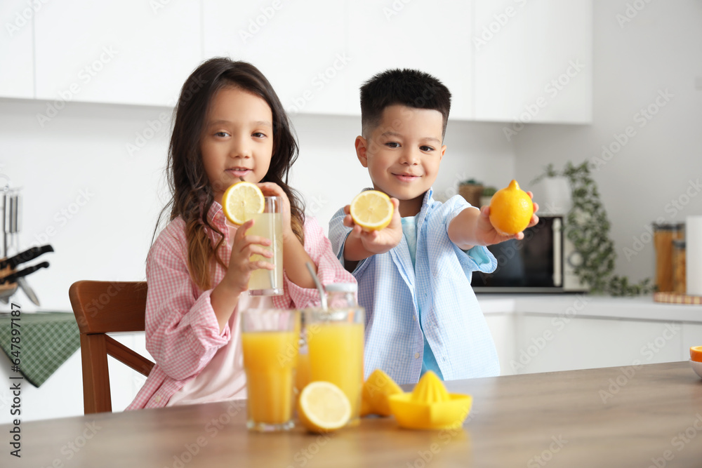 Little Asian children with glasses of fresh citrus juice and lemon slices sitting at table in kitche