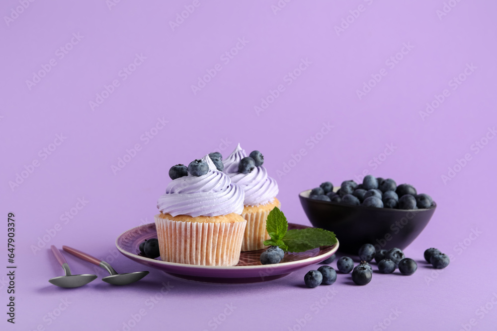 Plate of tasty cupcakes with blueberries on lilac background