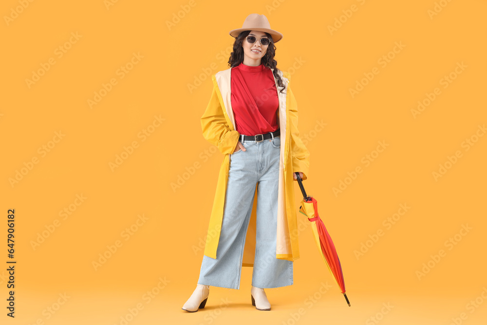 Stylish young Asian woman in fall clothes with umbrella on yellow background