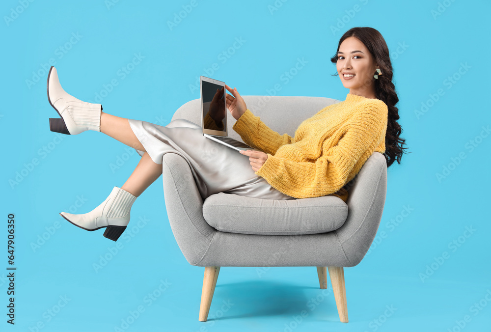 Young Asian woman using laptop in armchair on blue background