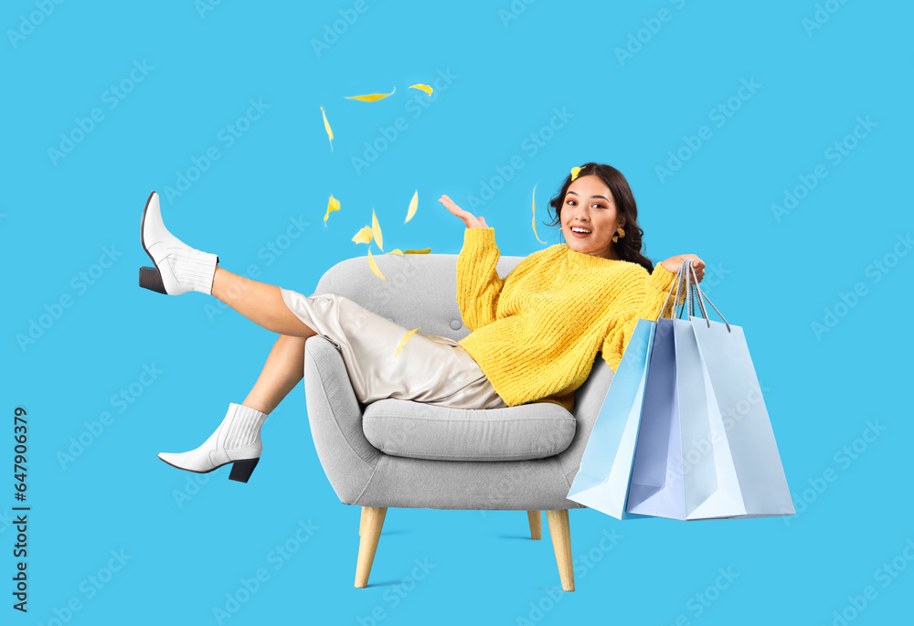 Young Asian woman in armchair with shopping bags and fallen leaves on blue background