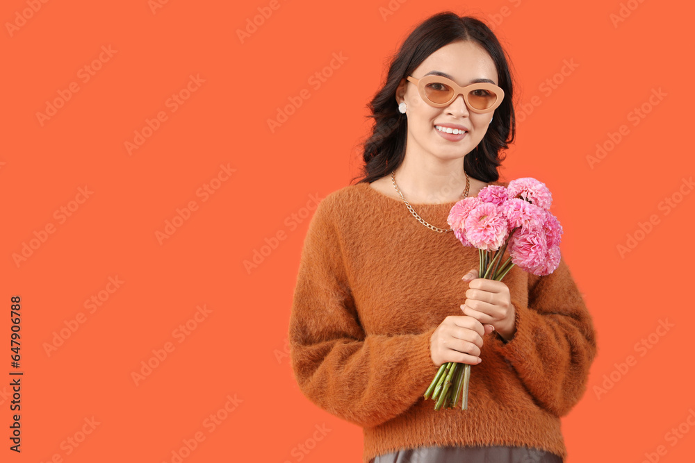 Stylish young Asian woman in sunglasses with flowers on orange background