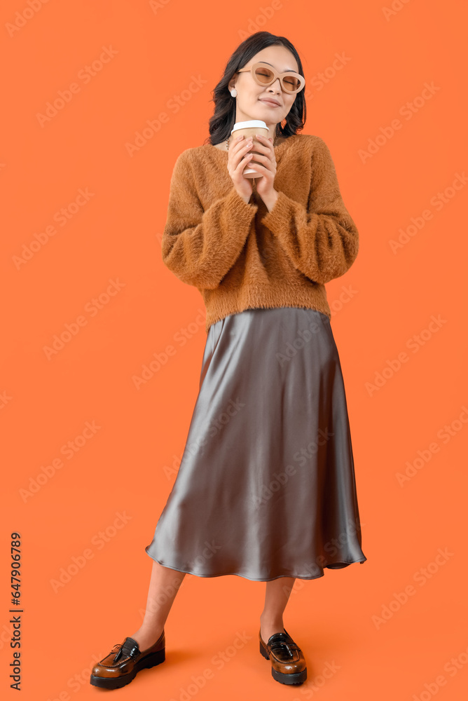 Stylish young Asian woman in fall clothes with coffee cup on orange background