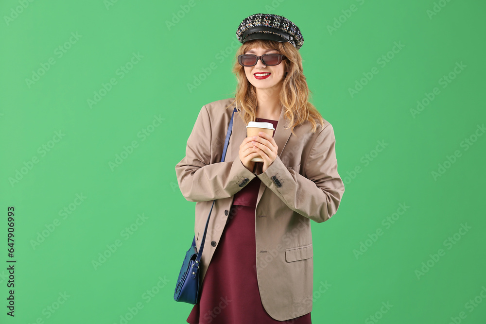 Stylish young woman in fall clothes with coffee cup on green background