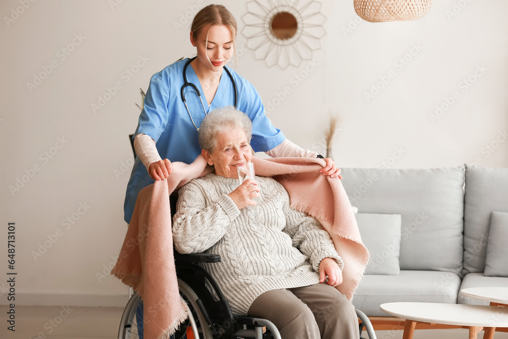 Young caregiver covering senior woman in wheelchair with plaid at home