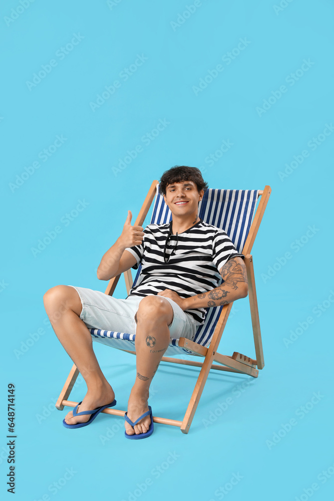 Young man showing thumb-up in deck chair on blue background