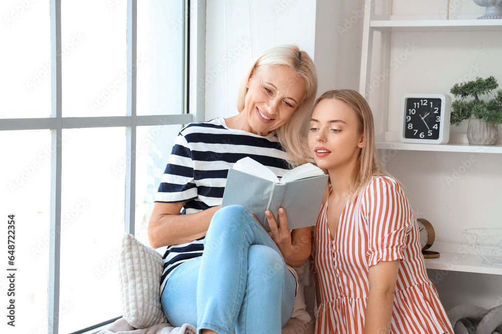 Happy young beautiful woman with her mother reading book together at home