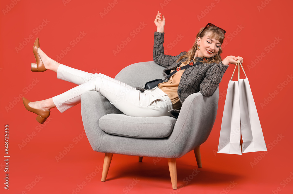 Stylish young woman with shopping bags in armchair on red background