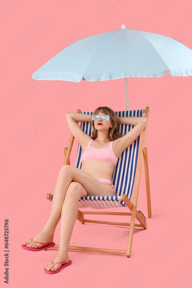 Young woman in deck chair with beach umbrella on pink background