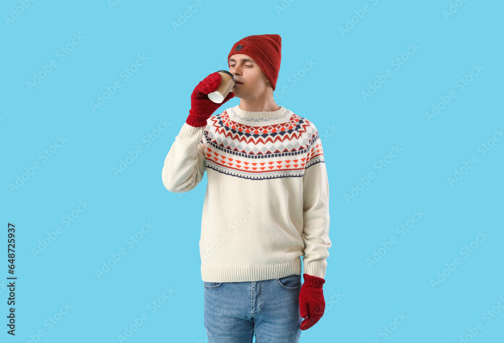 Young man in winter clothes drinking coffee on blue background