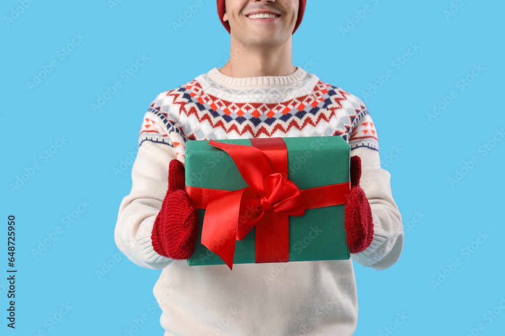 Young man in winter clothes with Christmas gift on blue background, closeup